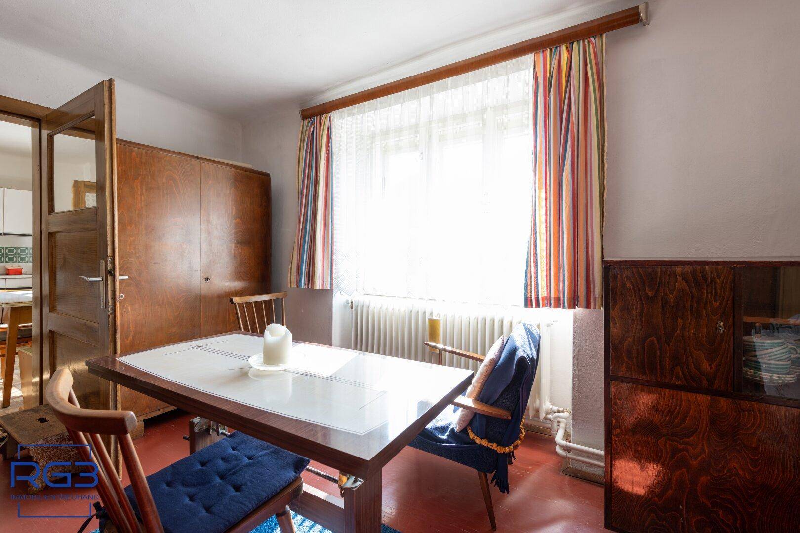 Esszimmer mit Holztisch, blauen Stühlen und bunten Vorhängen, durchflutet von Tageslicht.