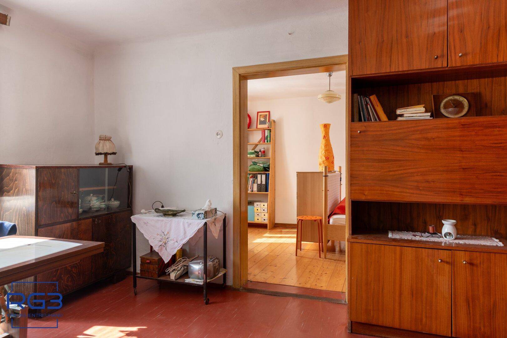 Wohnbereich mit Holzschrankwand und Durchgang zu einem hellen Schlafzimmer mit Holzboden.