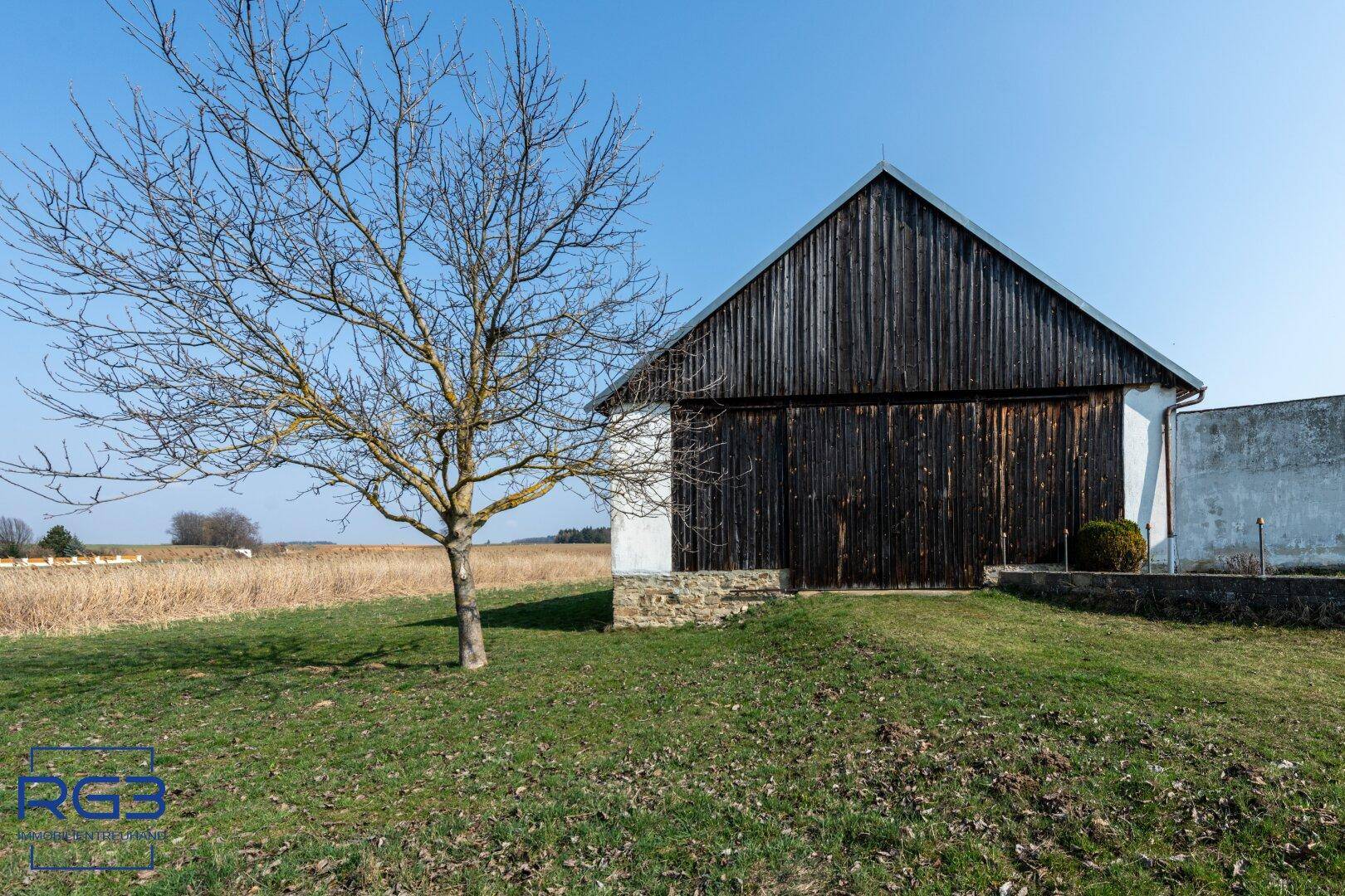 Rustikale Scheune mit Holzwänden und Ziegeldach, umgeben von einer weiten Wiesenlandschaft.
