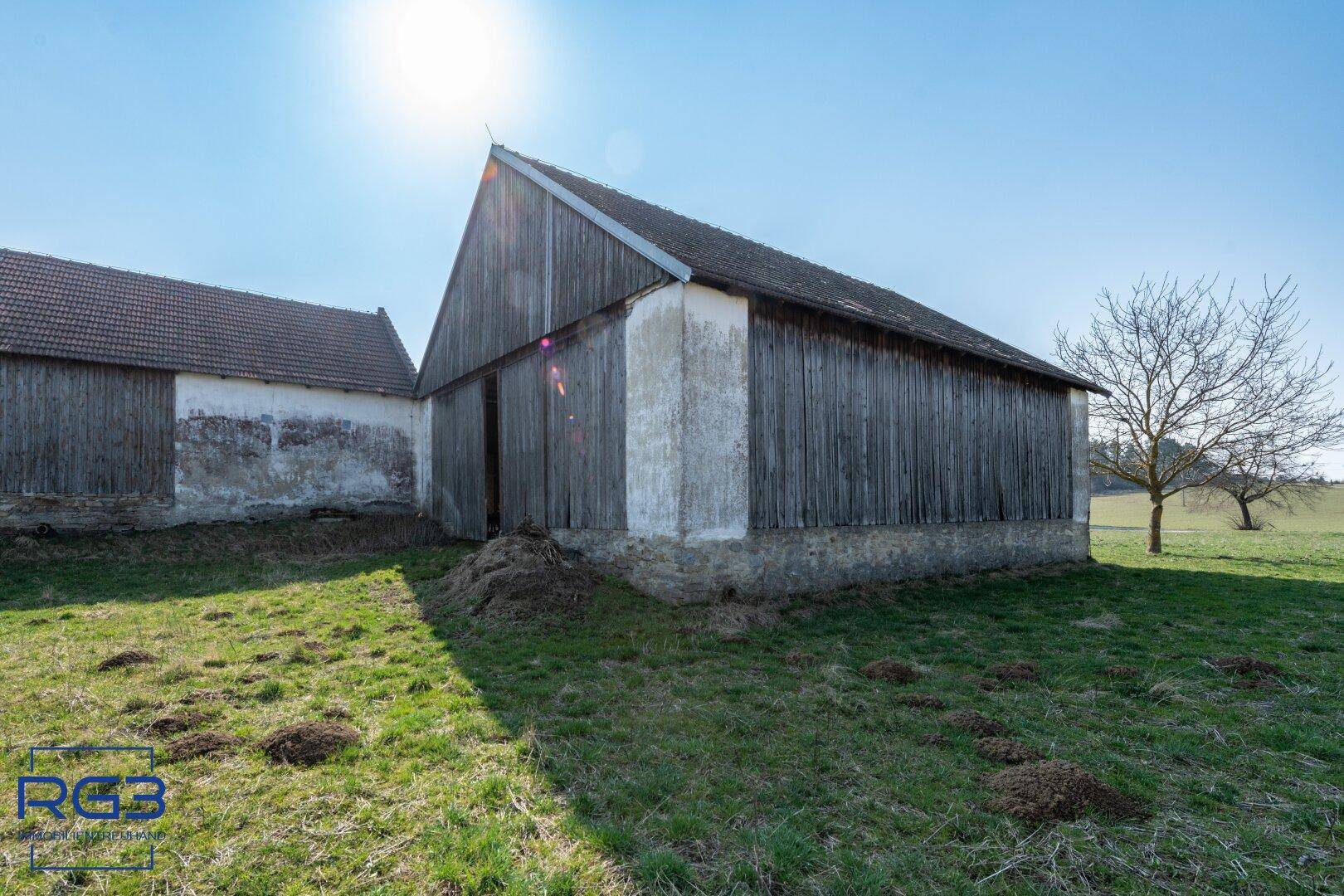 Große Holzscheune mit Ziegeldach und angrenzender Wiese in sonniger ländlicher Umgebung.
