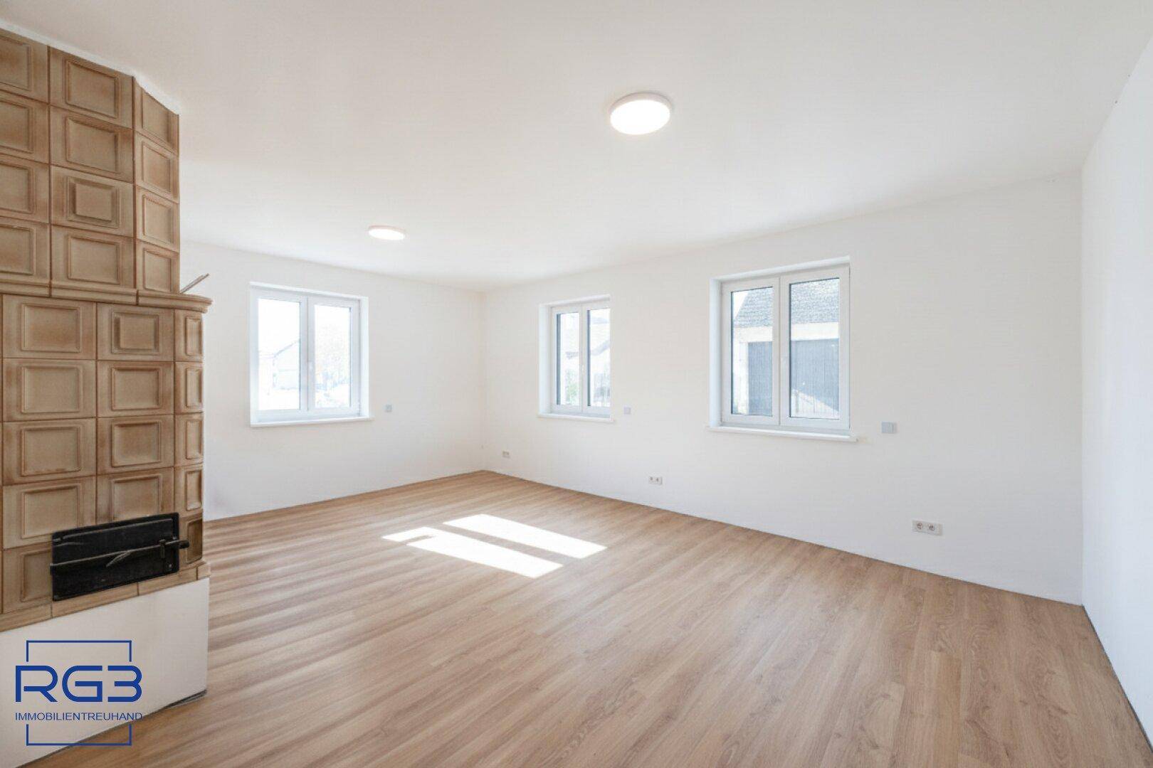 Geräumiges Wohnzimmer mit Kachelofen, Laminatboden und viel Tageslicht durch die Fenster.