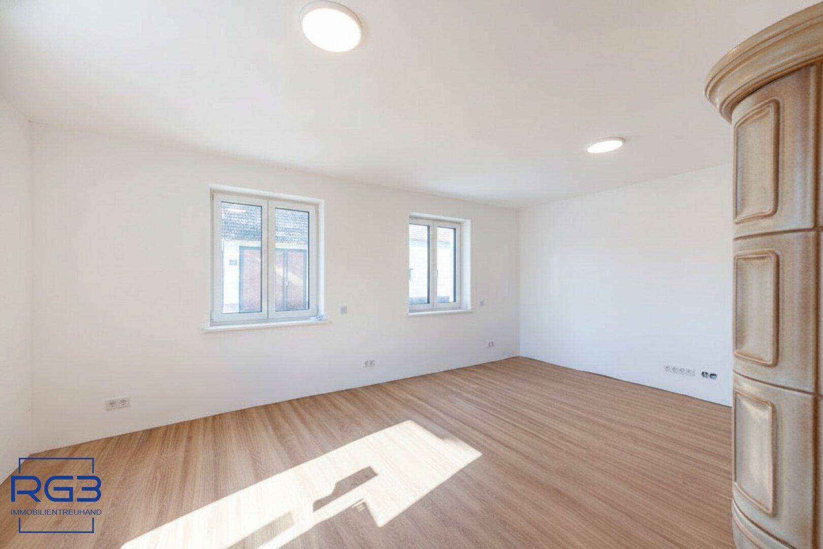 Helles Wohnzimmer mit traditionellem Kachelofen, Laminatboden und zwei Fenstern.
