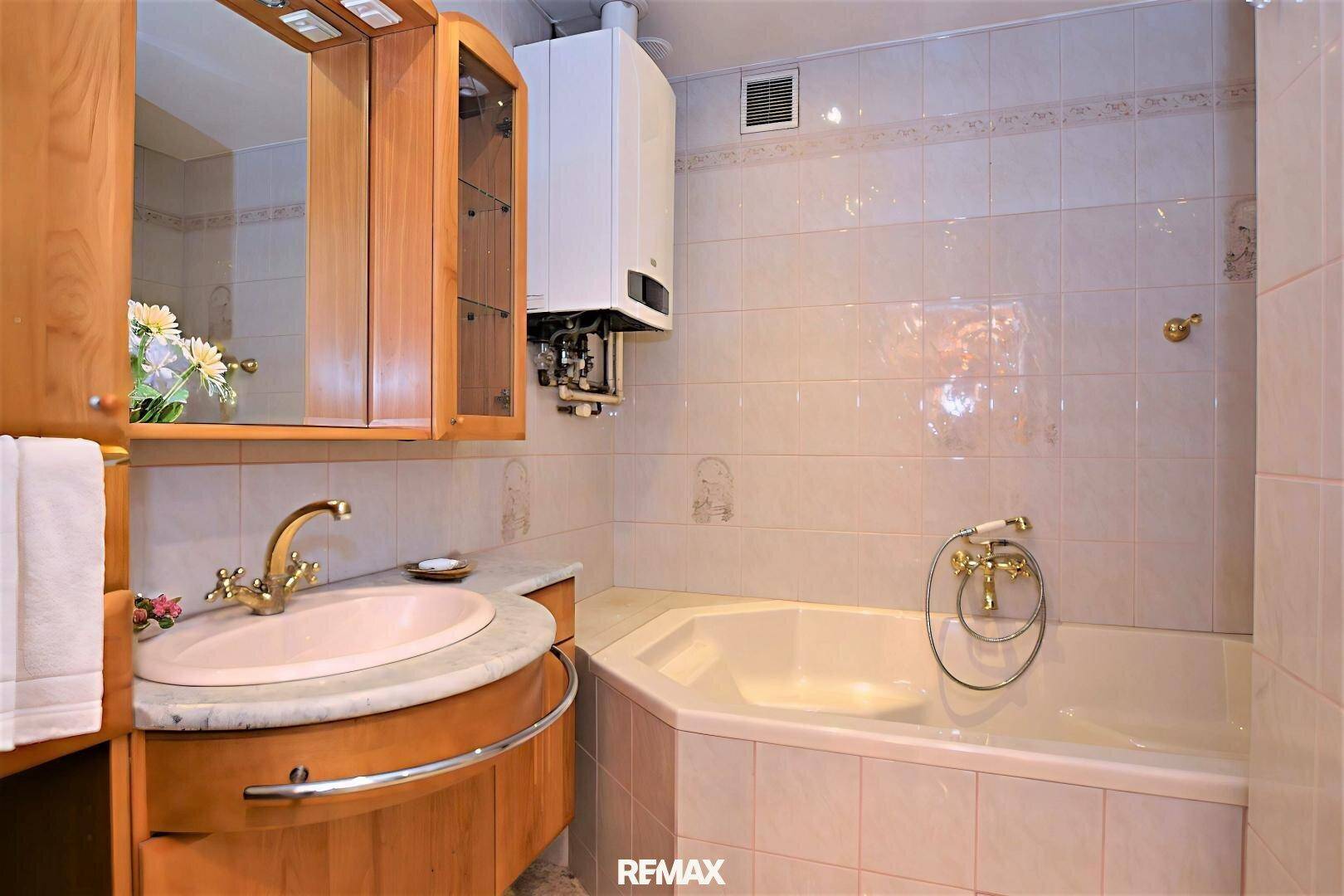 Badezimmer mit Eckbadewanne, Holzwaschtisch und gefliesten Wänden, ausgestattet mit einem Warmwasserbereiter.