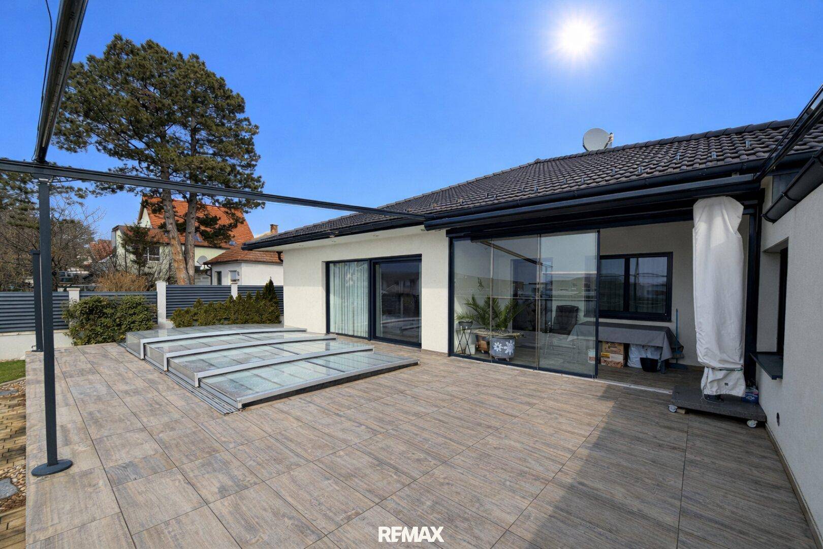Sonniger Außenbereich mit großer Terrasse, Rasenfläche und überdachtem Poolbereich am Haus.