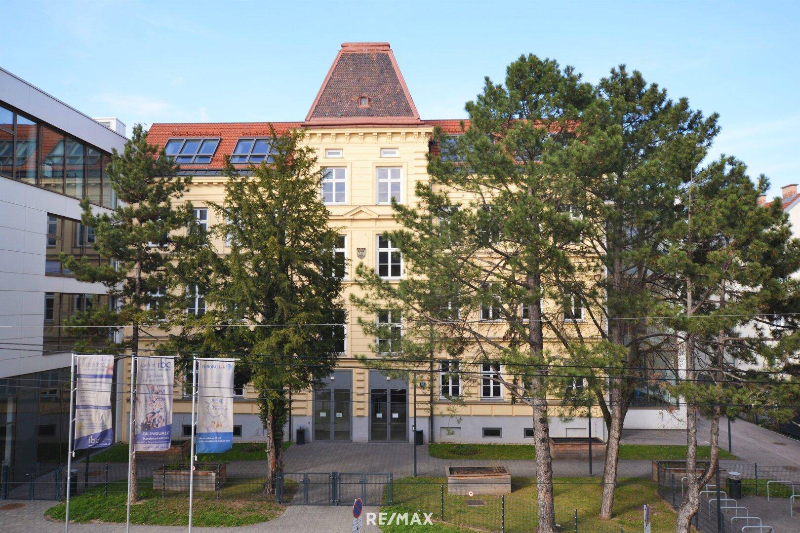 Historisches Gebäude mit gelber Fassade, roten Dachziegeln und hohen Bäumen davor.