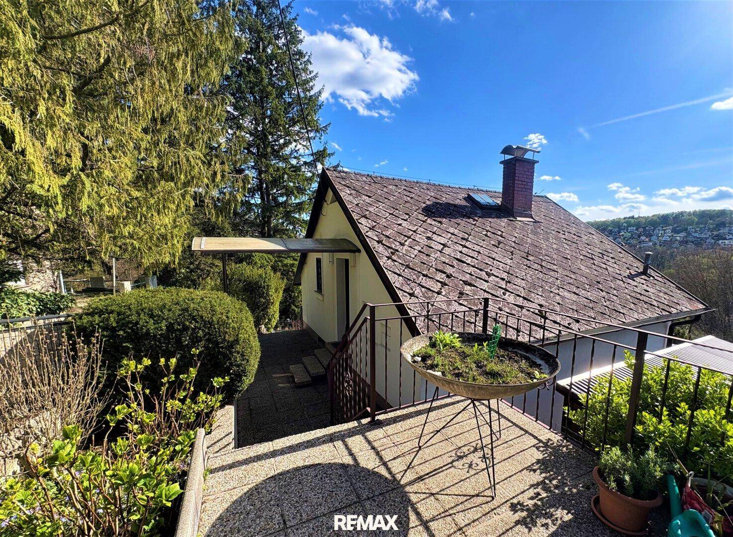 Seitlicher Blick auf das Haus mit Satteldach und einer Terrasse, umgeben von Bäumen und Sträuchern.