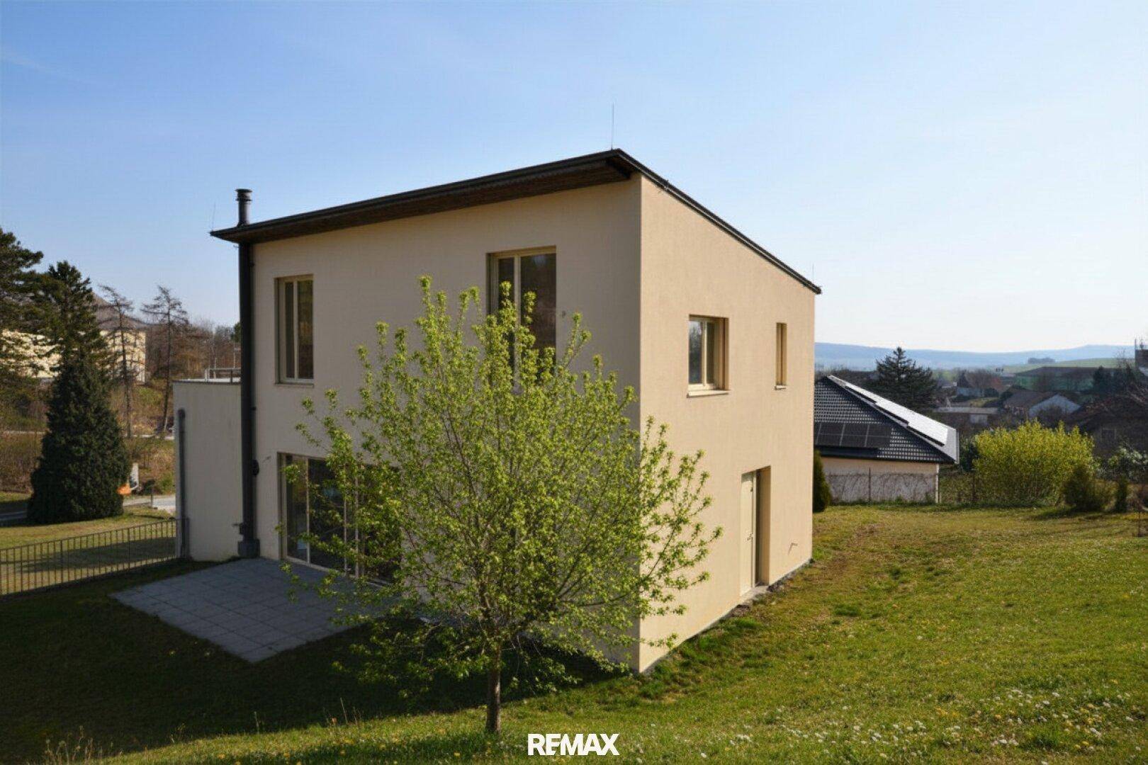 Modernes Einfamilienhaus mit heller Fassade, großen Fenstern und umliegender Grünfläche.