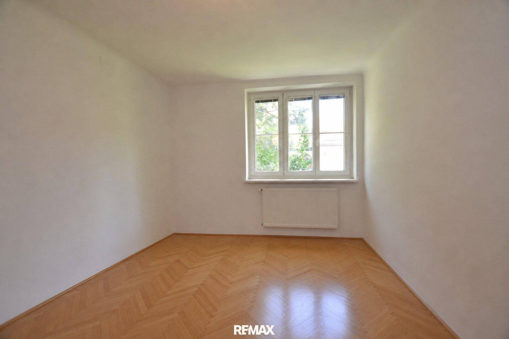 Leeres Schlafzimmer mit Fischgrätparkett und großem Fenster, bietet viel natürliches Licht.