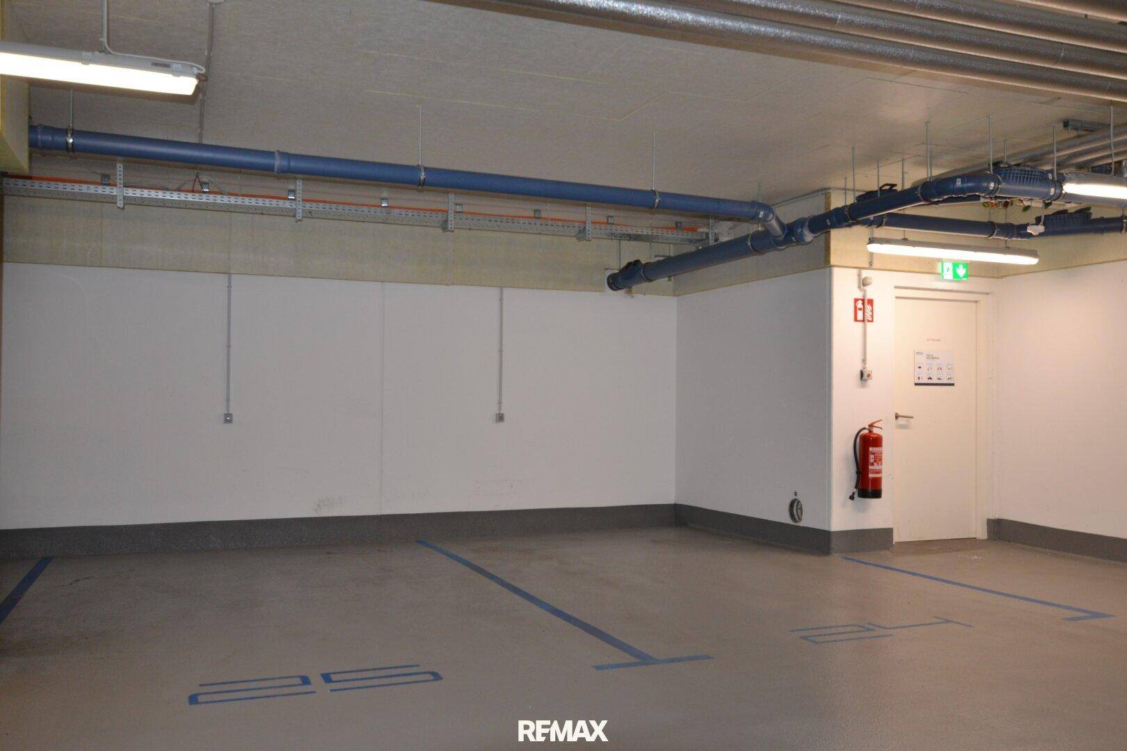 Tiefgarage mit markierten Stellplätzen, sichtbaren Installationen und Notausgang in einem gepflegten Zustand.