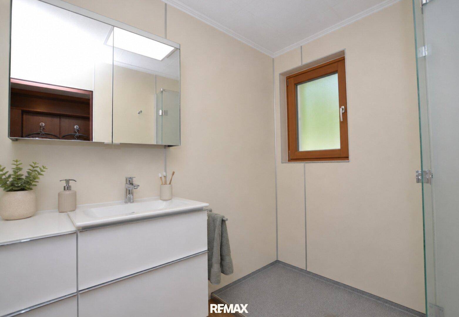 Modernes Badezimmer mit Einzelwaschbecken, Spiegelschrank, Duschbereich und Fenster für Tageslicht.