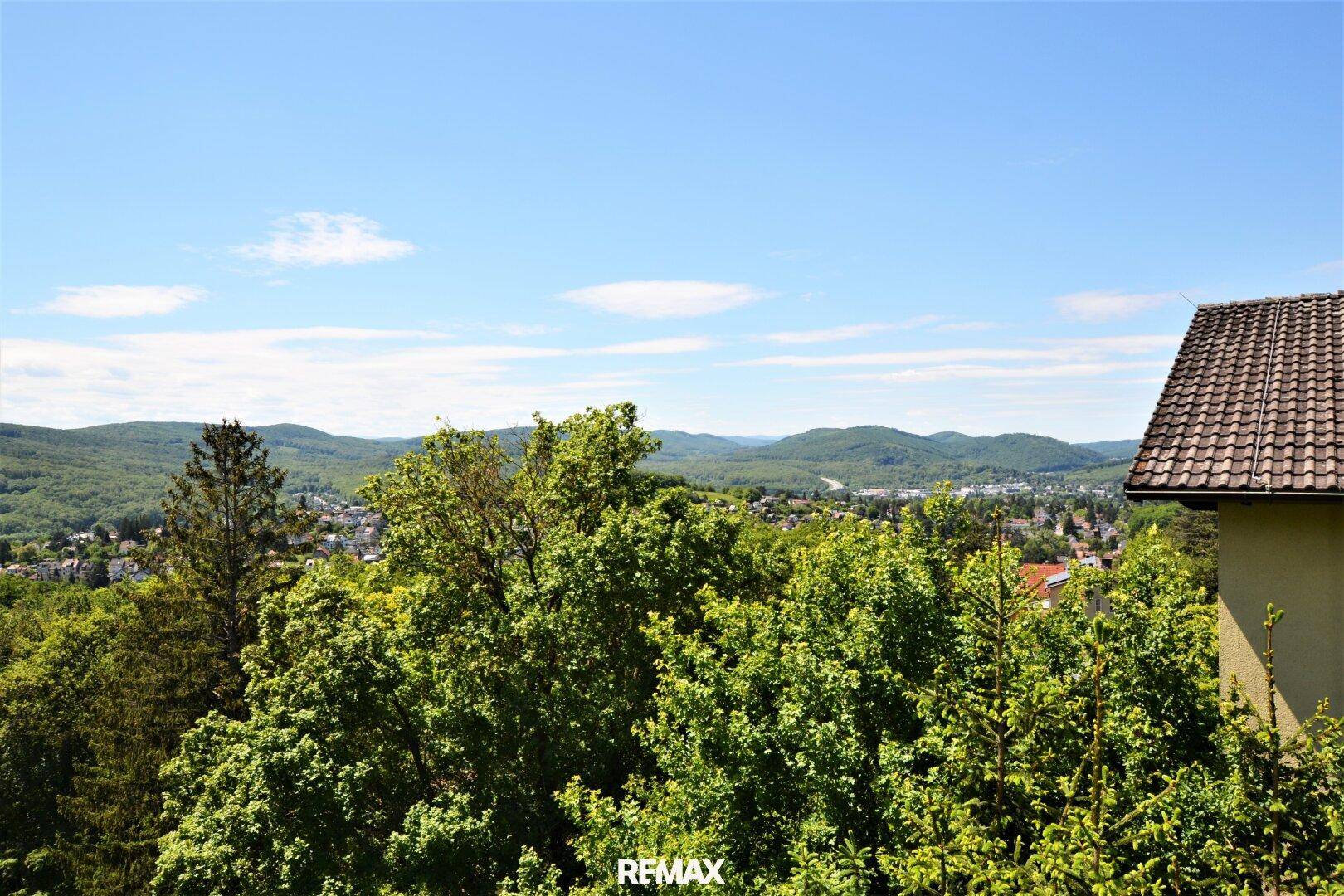 Weitläufiger Panoramablick über grüne Hügel und eine darunterliegende Ortschaft unter blauem Himmel.