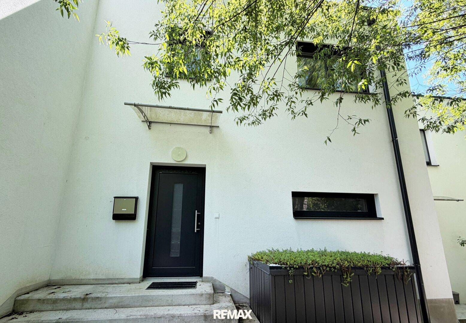 Moderner Hauseingang mit dunkler Tür und weißer Fassade, umgeben von Grün.