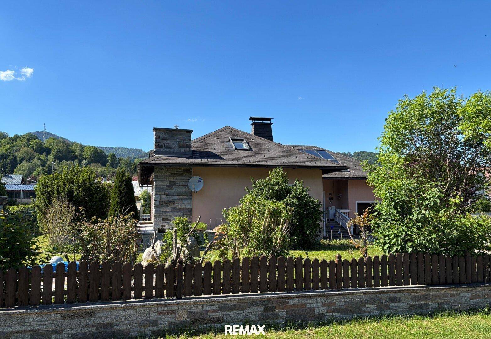 Rückansicht des Hauses mit Steinfassade, Schornstein und grüner Umgebung vor bewaldeten Hügeln.