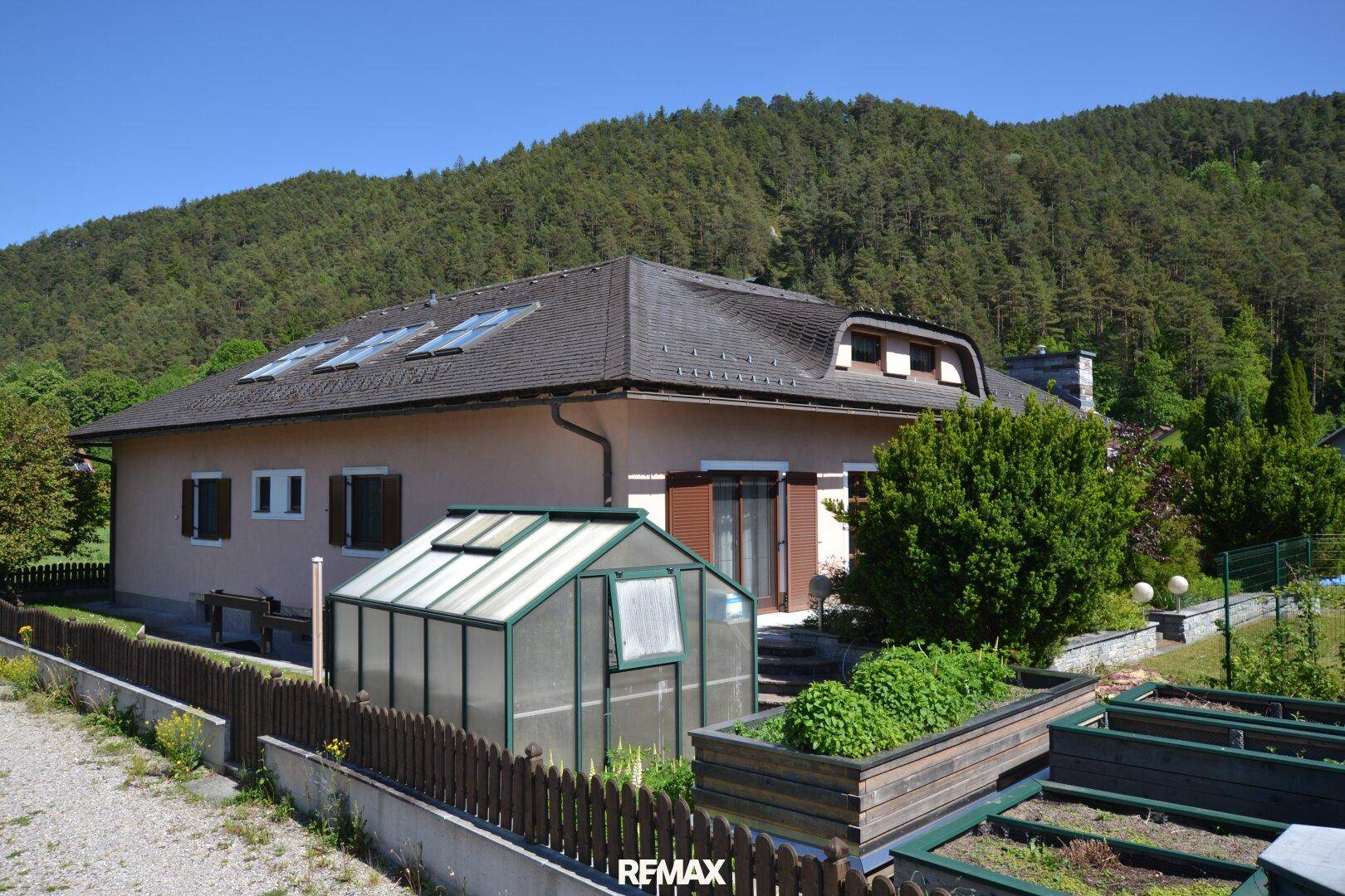 Seitenansicht des Hauses mit Gewächshaus, Hochbeeten und Blick auf die bewaldeten Berge.
