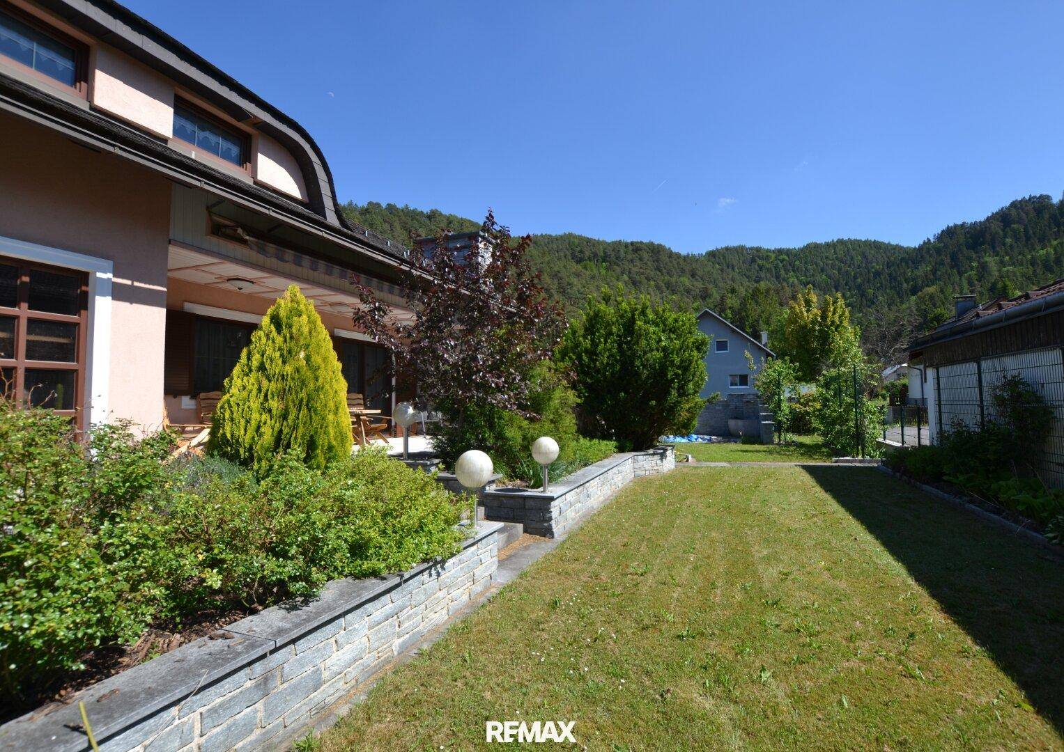 Seitlicher Gartenbereich mit Rasen, Sträuchern und Blick auf die bewaldeten Hügel.