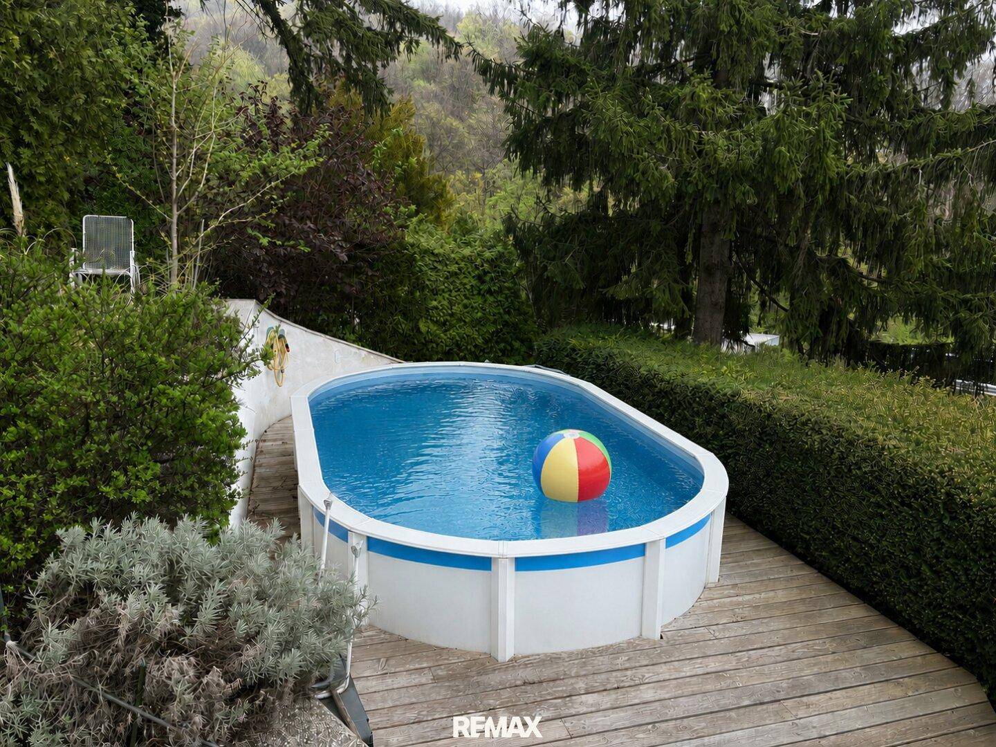 Ovaler Aufstellpool im Garten auf einer Holzterrasse, umgeben von Bäumen und Hecken.