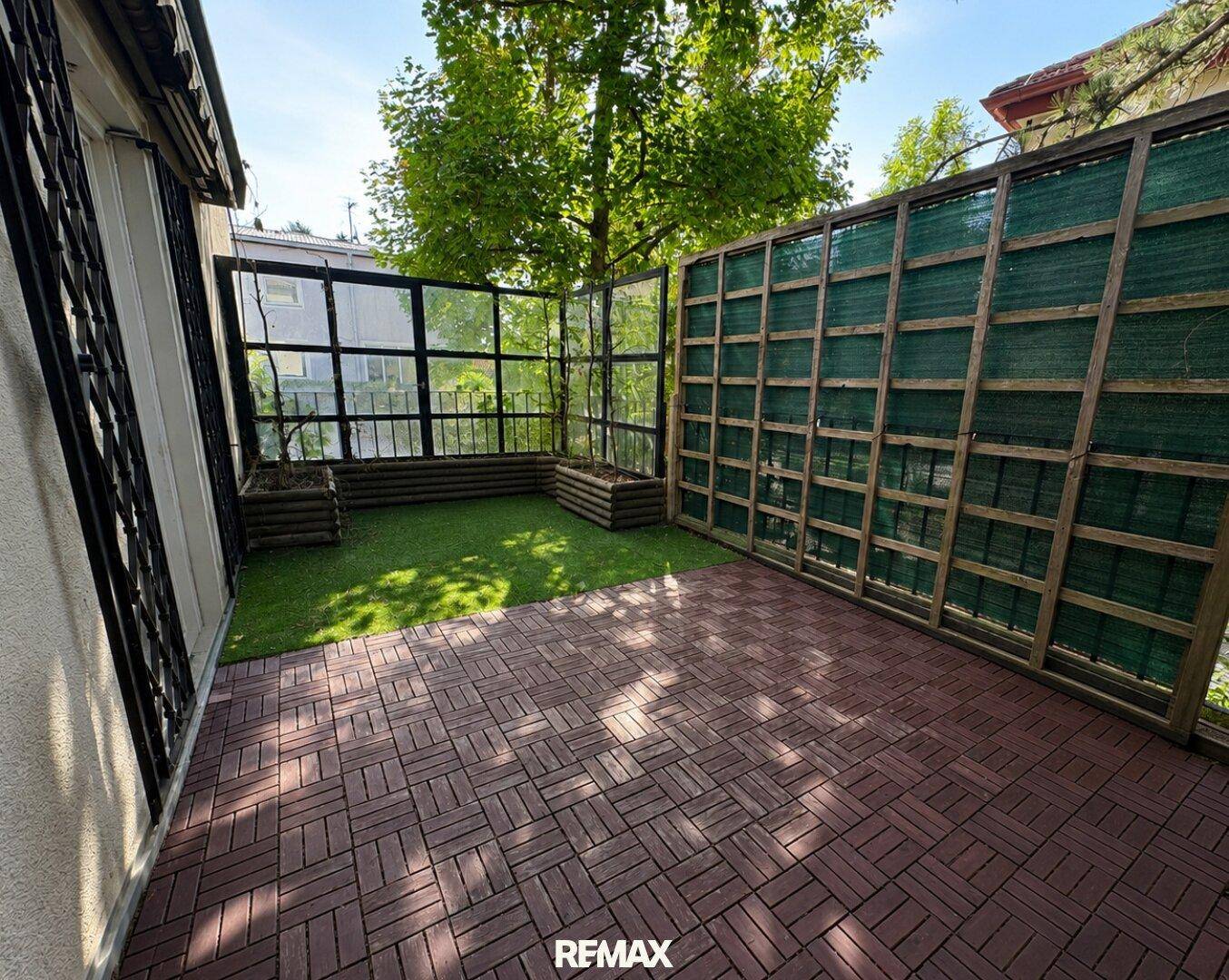 Kleine Terrasse mit Holzbodenfliesen und Kunstrasen, umgeben von Sichtschutz.