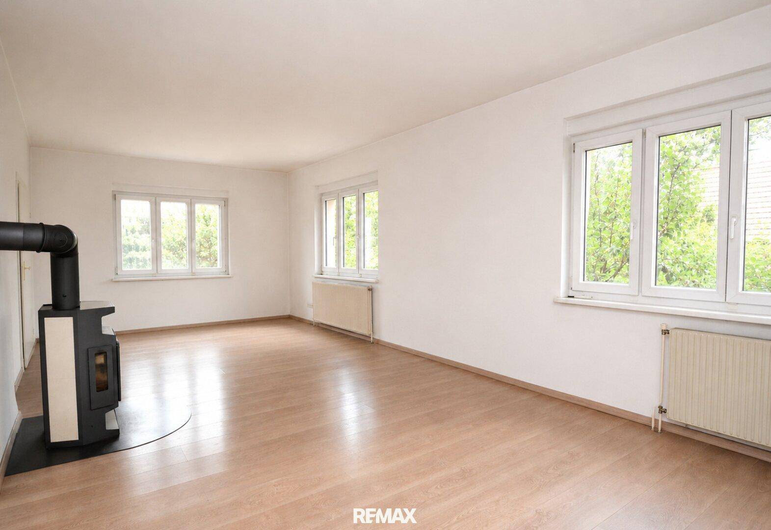 Geräumiges, helles Wohnzimmer mit Holzboden, großen Fenstern und einem modernen Kaminofen.