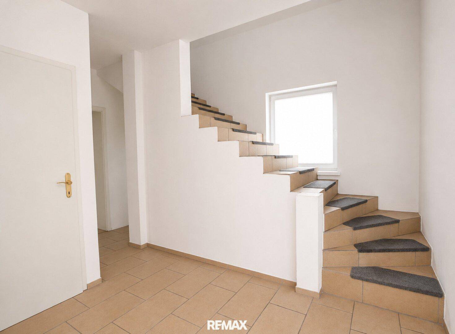 Heller Flur mit Treppe zum Obergeschoss und Fliesenboden, gut beleuchtet durch ein Fenster.