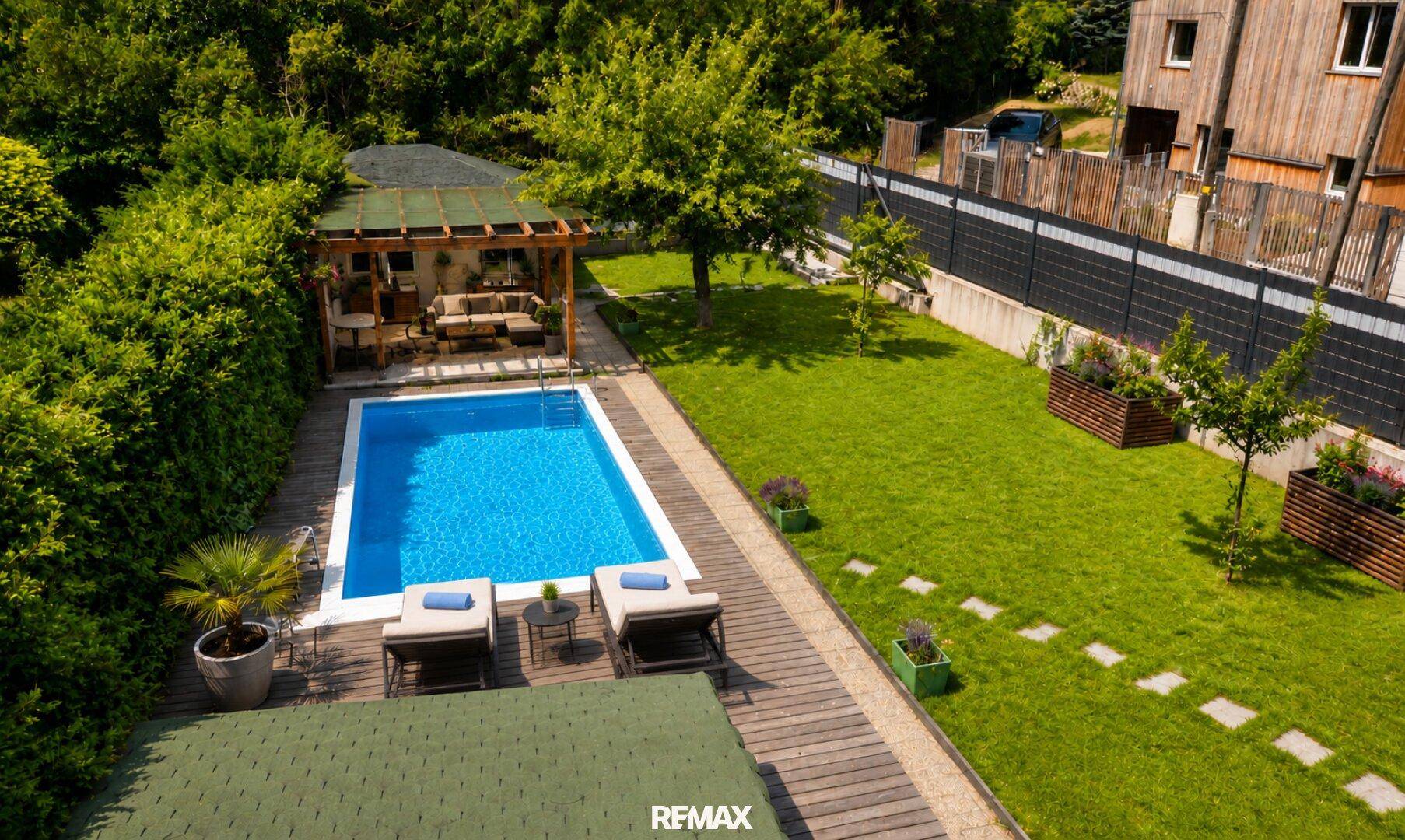 Vogelperspektive des Gartens mit Pool, Holzdeck, Liegestühlen und angrenzendem Rasenbereich.