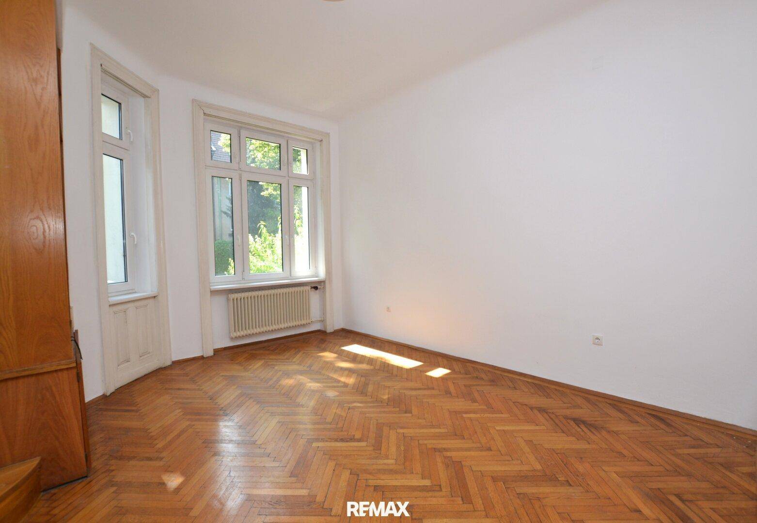 Helles Zimmer mit Fischgrätparkett, großen Fenstern und Heizkörper unter dem Fenster.