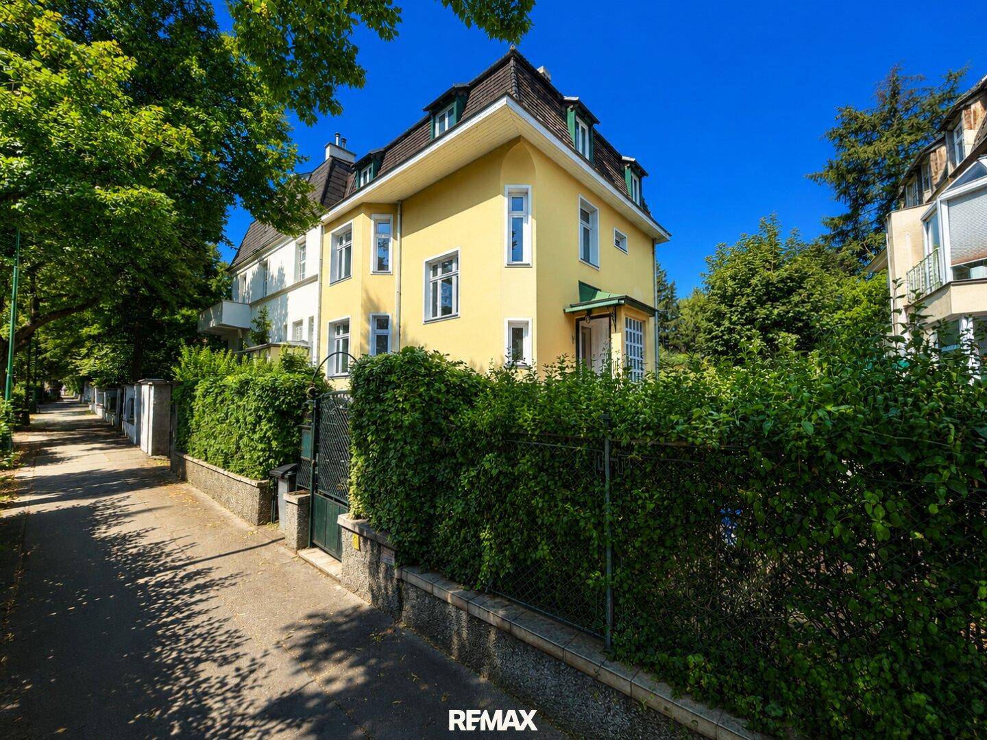 Historische Villa mit gelber Fassade und üppiger Hecke entlang der ruhigen Straße.