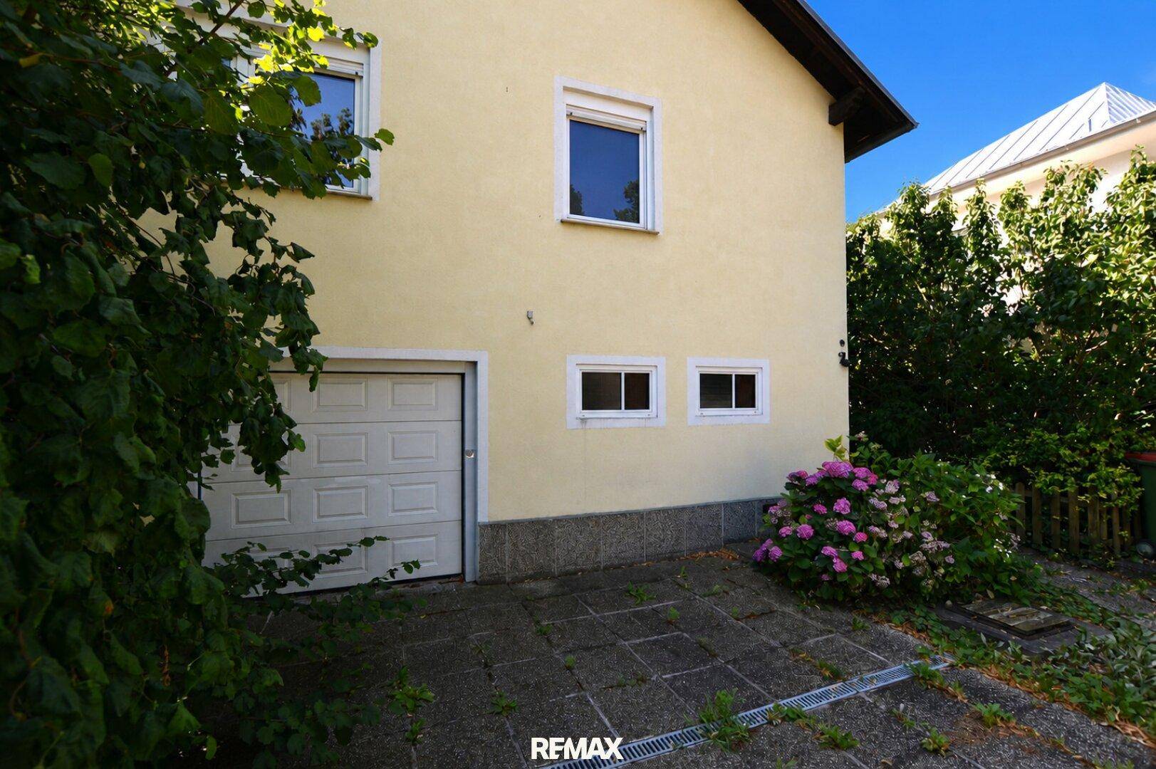 Seitenansicht des Hauses mit Garagentor, gepflasterter Zufahrt und blühenden Hortensien.