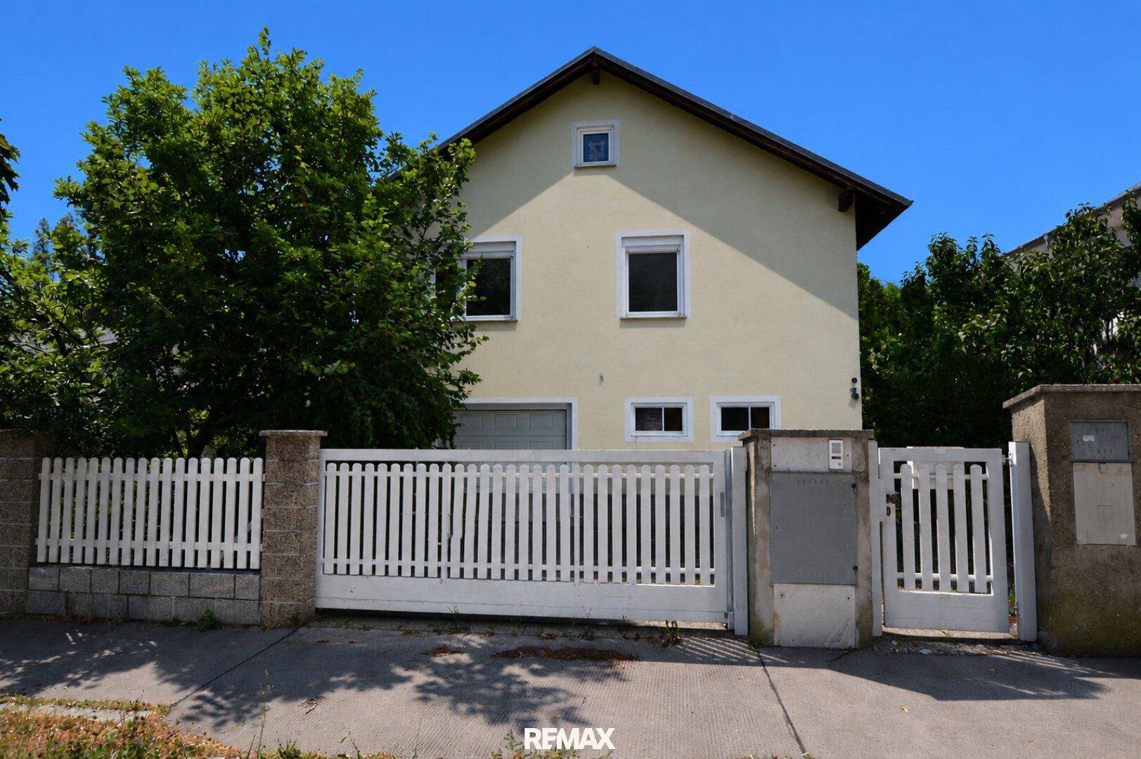 Vorderansicht des Hauses mit weißem Zaun, Einfahrt und Rollläden an den Fenstern.