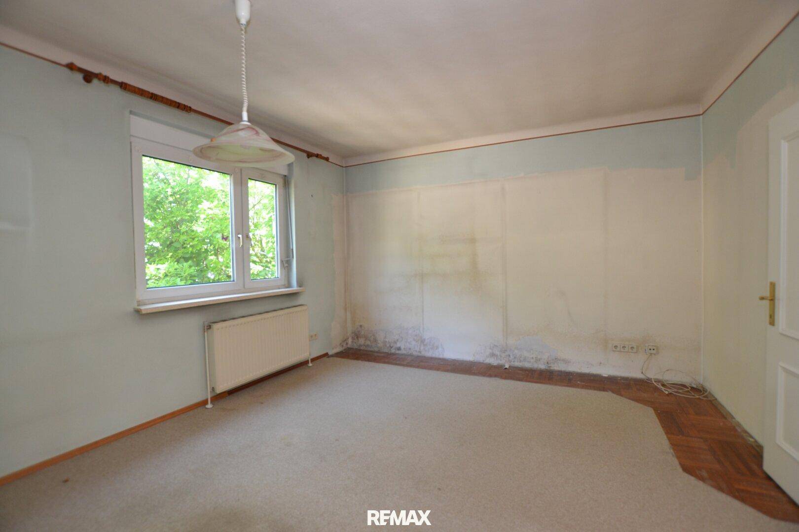 Schlafzimmer in ursprünglichem Zustand mit Teppichboden und Fensterblick ins Grüne.