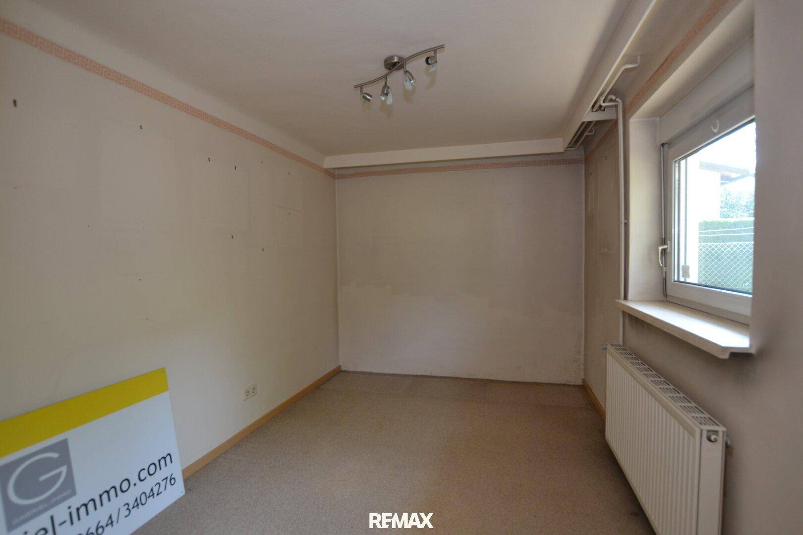 Zimmer in ursprünglichem Zustand mit Teppichboden und Fensterblick ins Grüne.