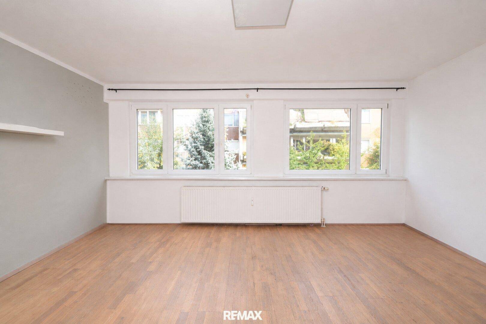 Geräumiges, unmöbliertes Wohnzimmer mit großen Fenstern und hellem Parkettboden.