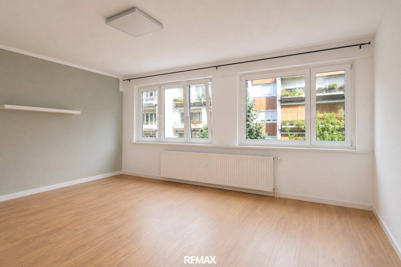 Leeres Wohnzimmer mit Parkettboden und Blick auf die umliegenden Gebäude und Bäume.