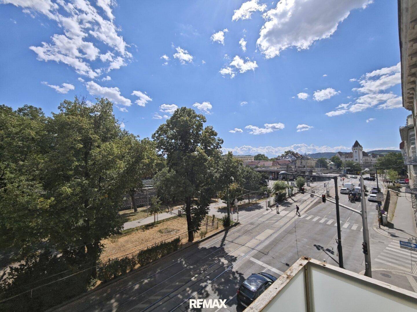 Weitblick über eine belebte Straße mit Bäumen und städtischer Architektur unter blauem Himmel.