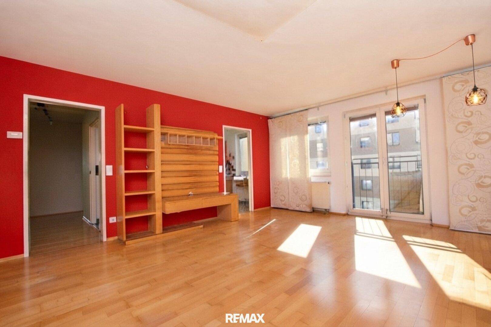 Geräumiges Wohnzimmer mit roter Wand, Parkettboden und viel Tageslicht.