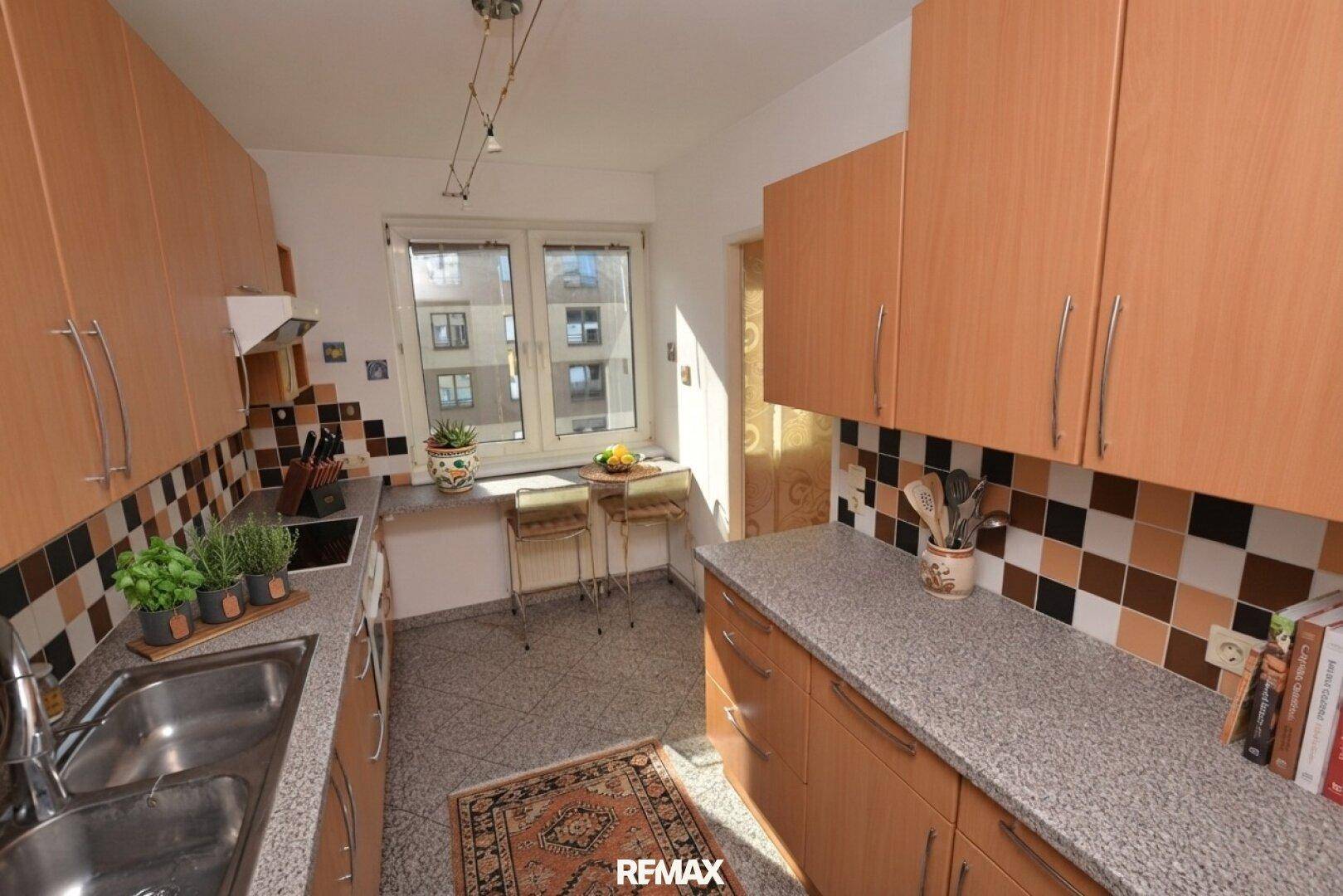 Funktionale Küche mit Holzschränken, Fliesenboden und Essbereich am Fenster.