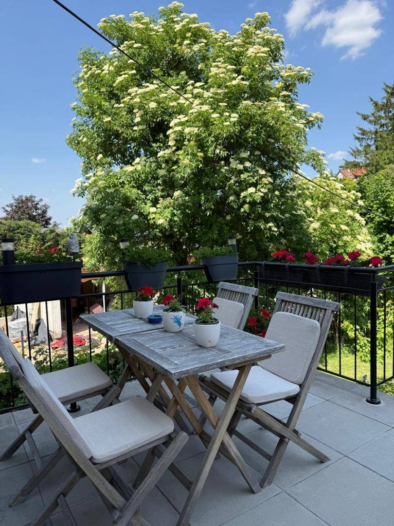 Sonniger Balkon mit Holztisch und Stühlen, umgeben von blühenden Bäumen und Pflanzen.