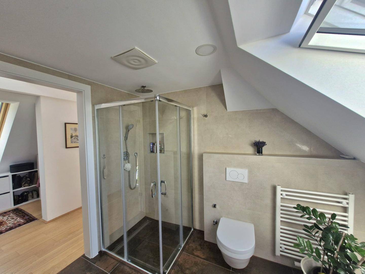 Modernes Badezimmer im Dachgeschoss mit Dusche, WC, Handtuchheizkörper und Dachfenster für Tageslicht.