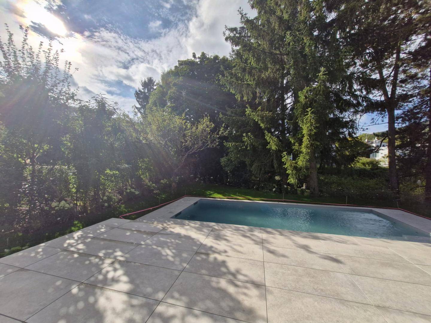 Sonniger Poolbereich mit großzügiger Terrasse und üppigem Grün, ideal zum Entspannen.