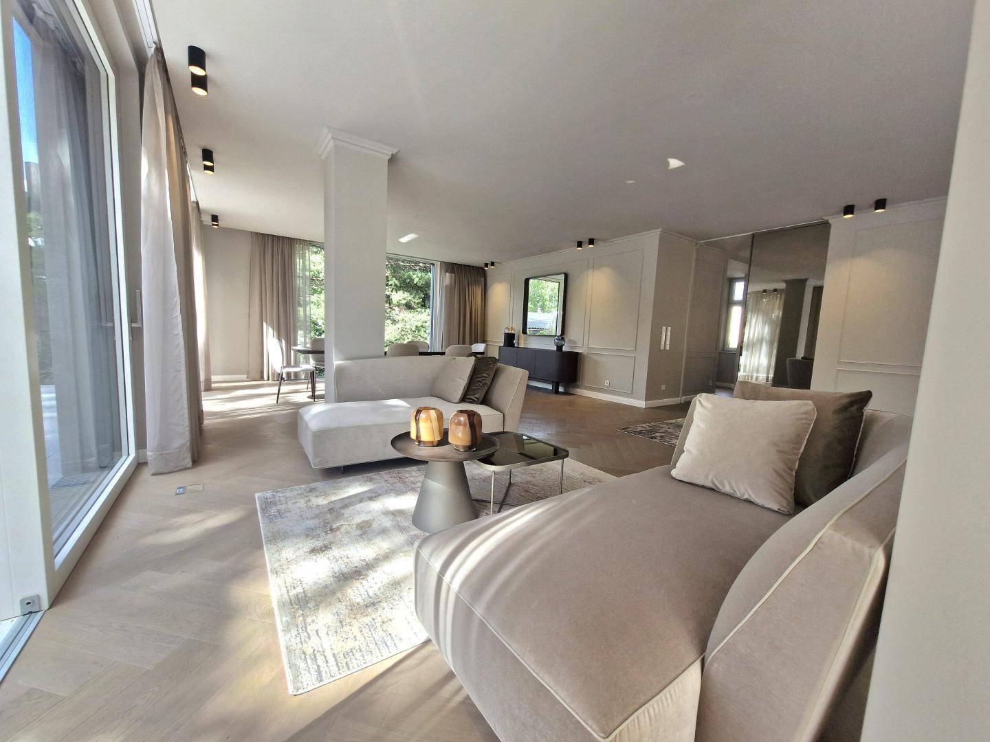 Lichtdurchfluteter Wohnbereich mit modernen Sofas, Fischgrätparkett und großen Fenstern zum Außenbereich.