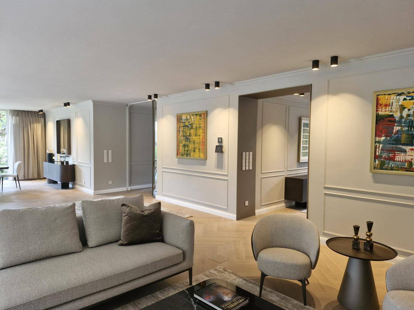 Geräumiges Wohnzimmer mit modernen Sofas, Kunstwerken und Fischgrätparkett, offen zu anderen Bereichen.