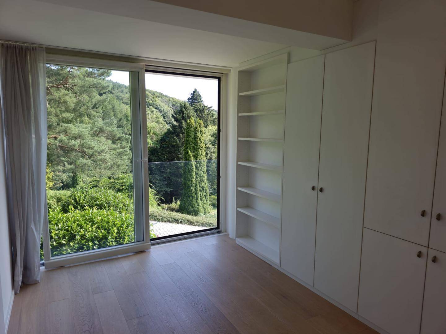 Schlafzimmer mit Holzboden, Einbauschränken und großem Fenster mit Blick auf die grüne Umgebung.