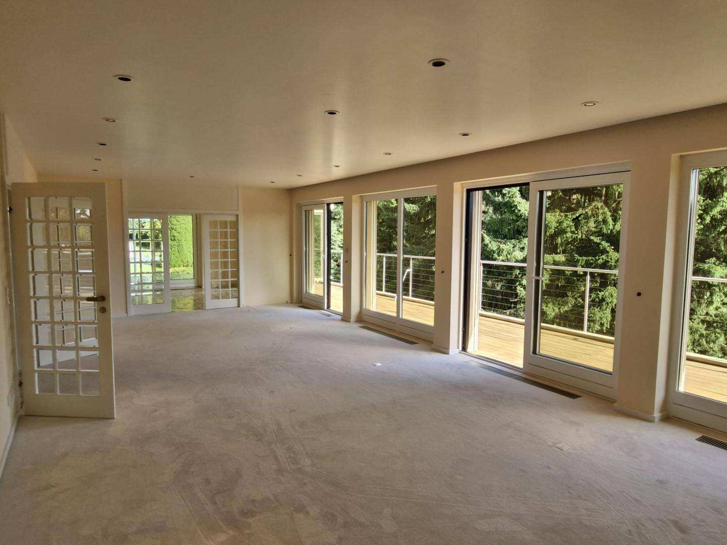 Geräumiger Wohnbereich mit vielen Fenstertüren, die Zugang zu einem Balkon und Blick ins Grüne bieten.