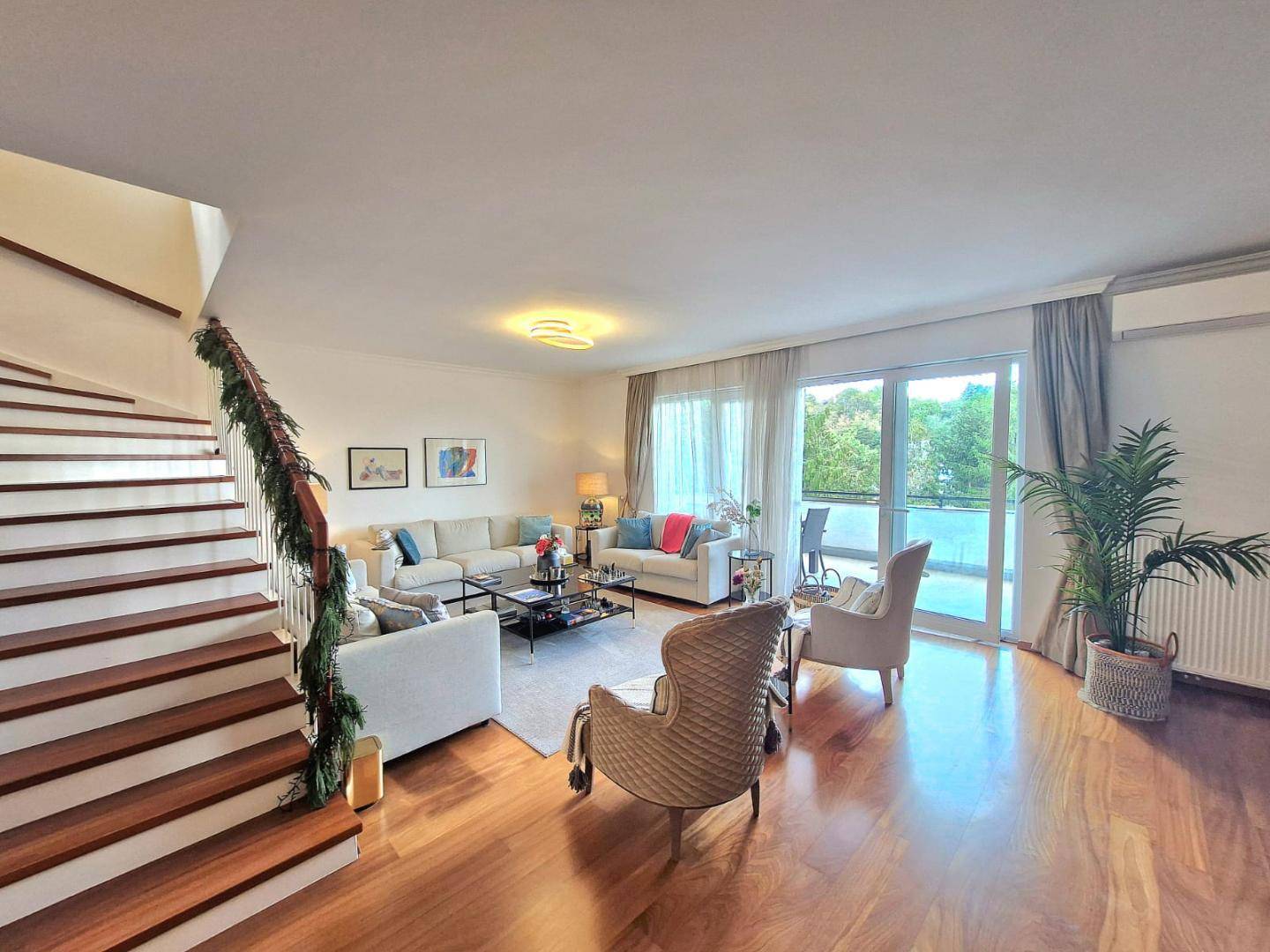 Helles Wohnzimmer mit Zugang zum Balkon, bequemen Sofas und einer eleganten Holztreppe.