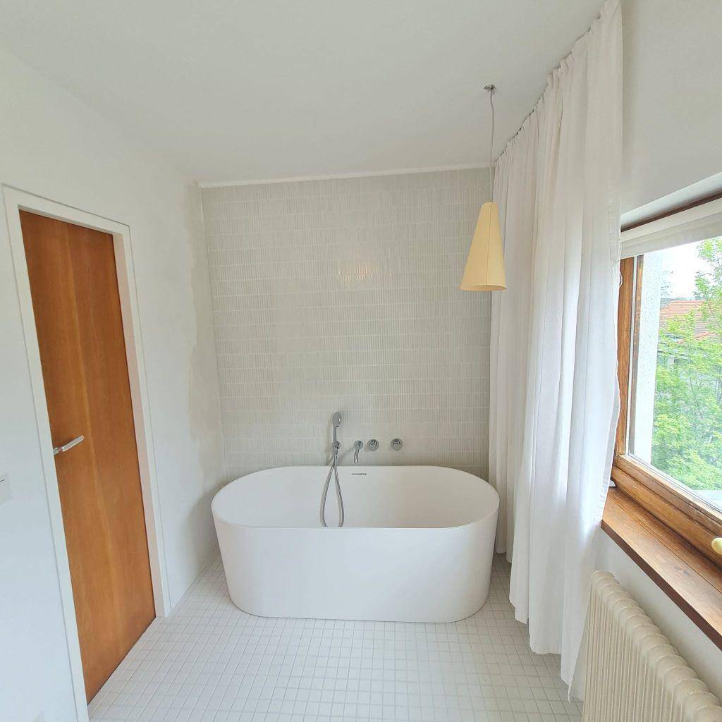 Helles Badezimmer mit freistehender Badewanne, weißen Fliesen und Fenster für Tageslicht.