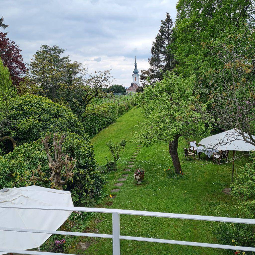 Panoramablick auf den weitläufigen Garten mit üppiger Vegetation und einem Kirchturm in der Ferne.