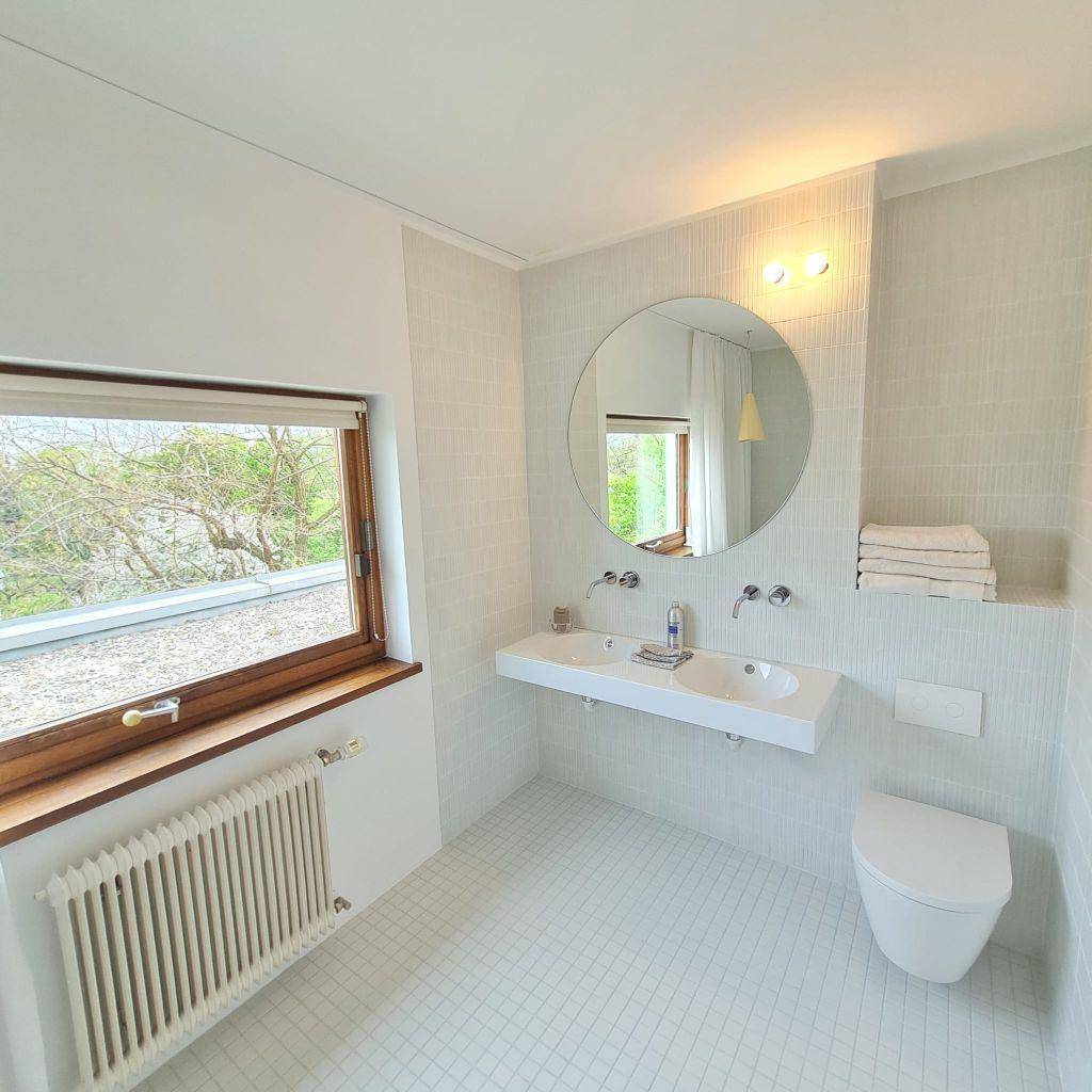 Modernes Badezimmer mit Doppelwaschbecken, Wand-WC und Fenster mit Blick ins Grüne.
