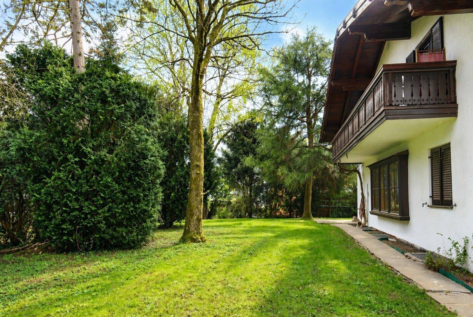 Blick auf die Hausfassade mit Holzbalkon und einem weitläufigen Gartenbereich mit altem Baumbestand.