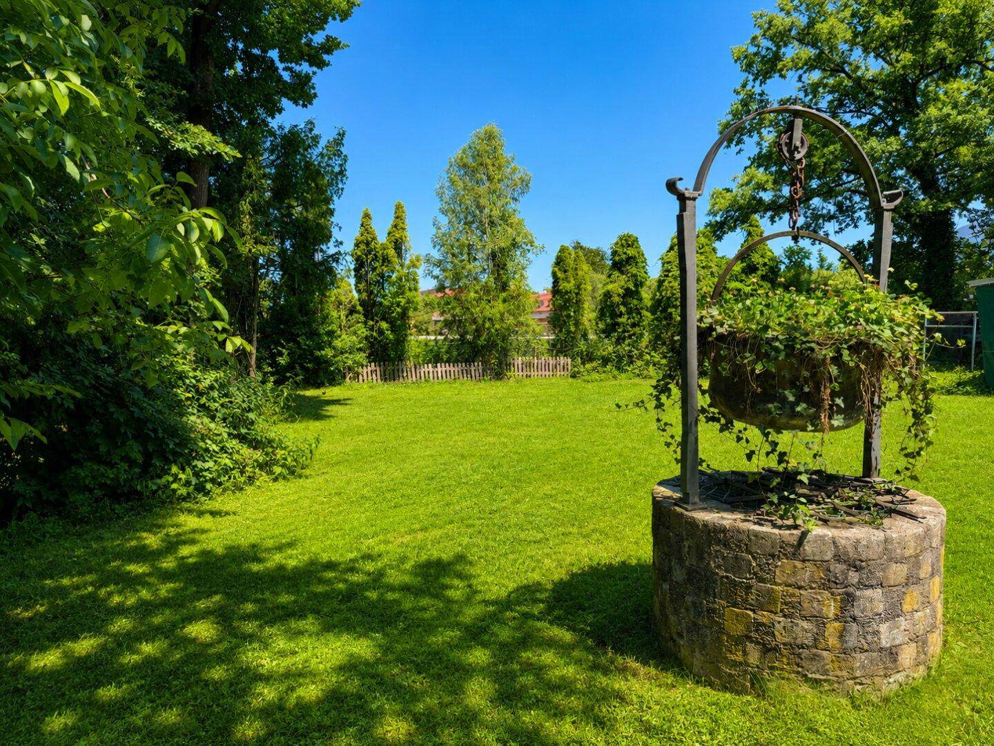 Historischer Steinbrunnen mit Pflanzen in einem weitläufigen, grünen Garten unter blauem Himmel.