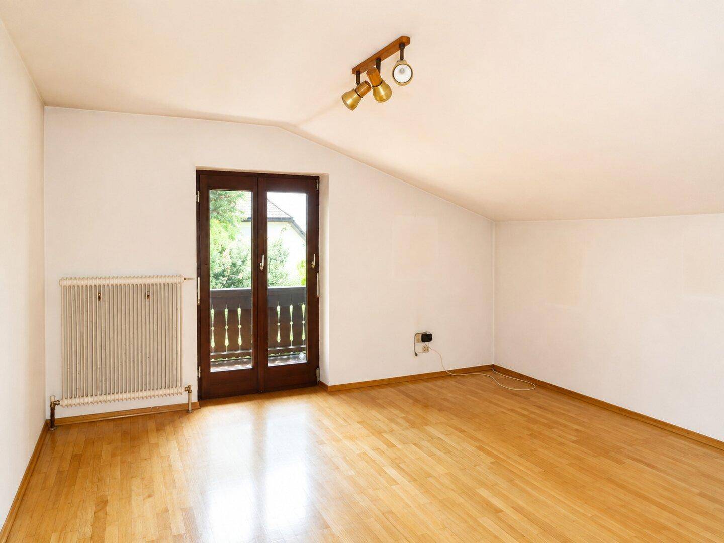 Helles Zimmer im Dachgeschoss mit Holzboden, Heizkörper und Zugang zu einem kleinen Balkon.