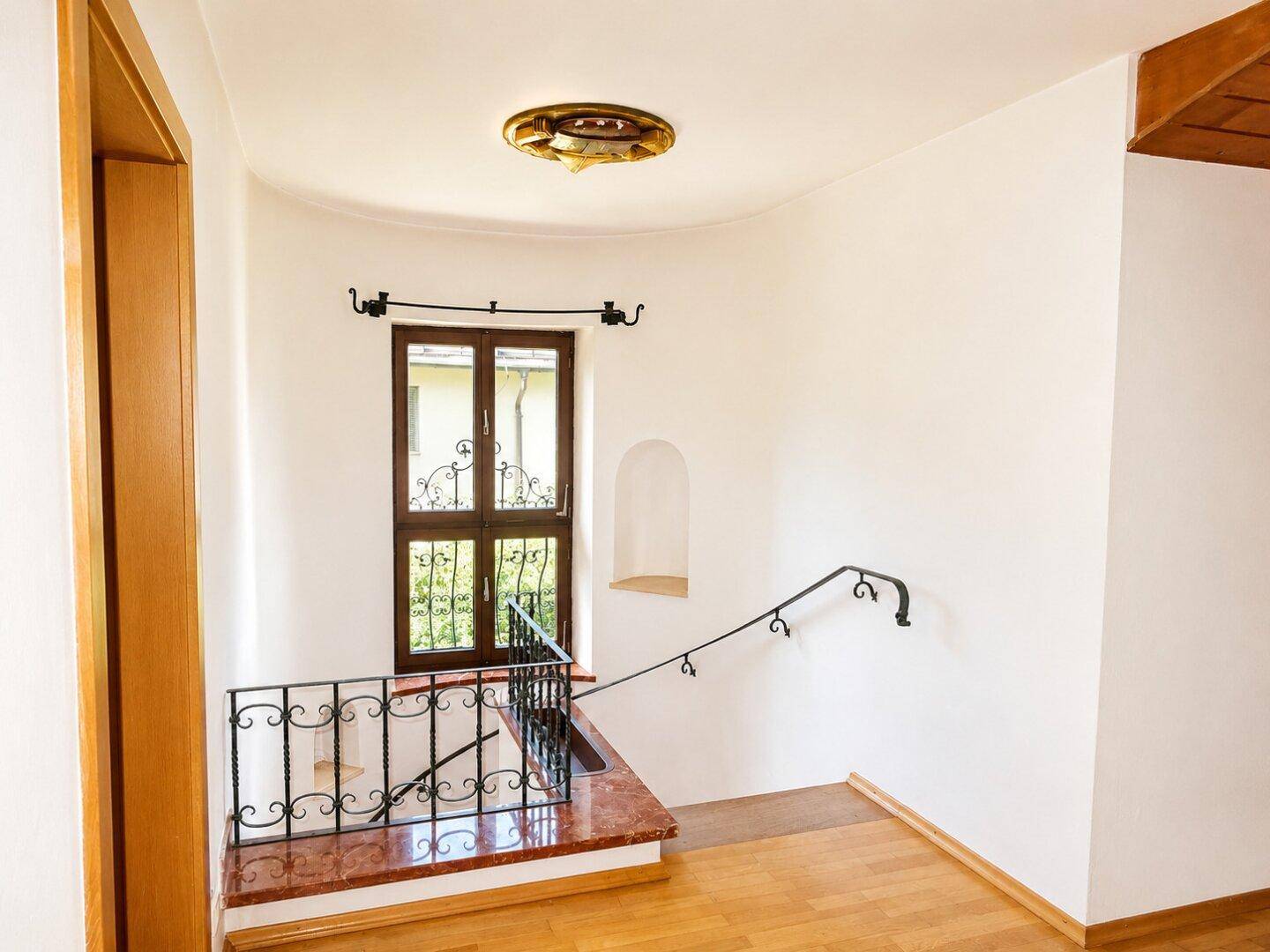 Heller Flurbereich mit Marmortreppe, schmiedeeisernem Geländer und Fenster mit Blick ins Grüne.